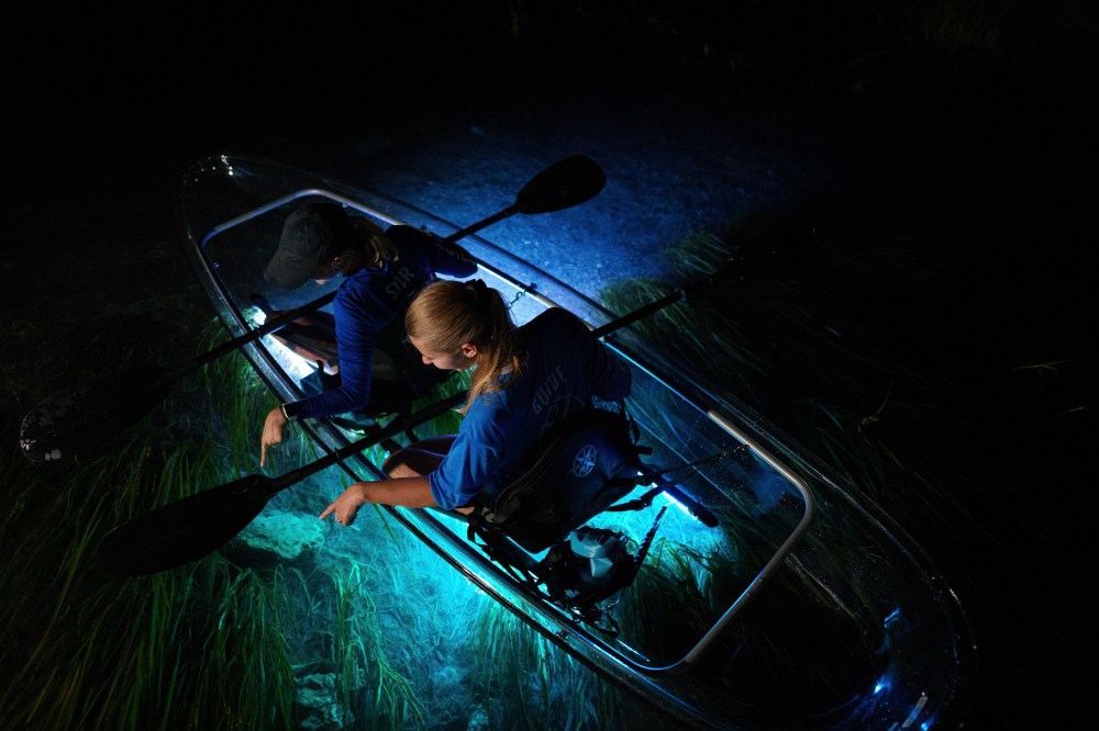 Two people in clear kayak with blue lights on dark water.