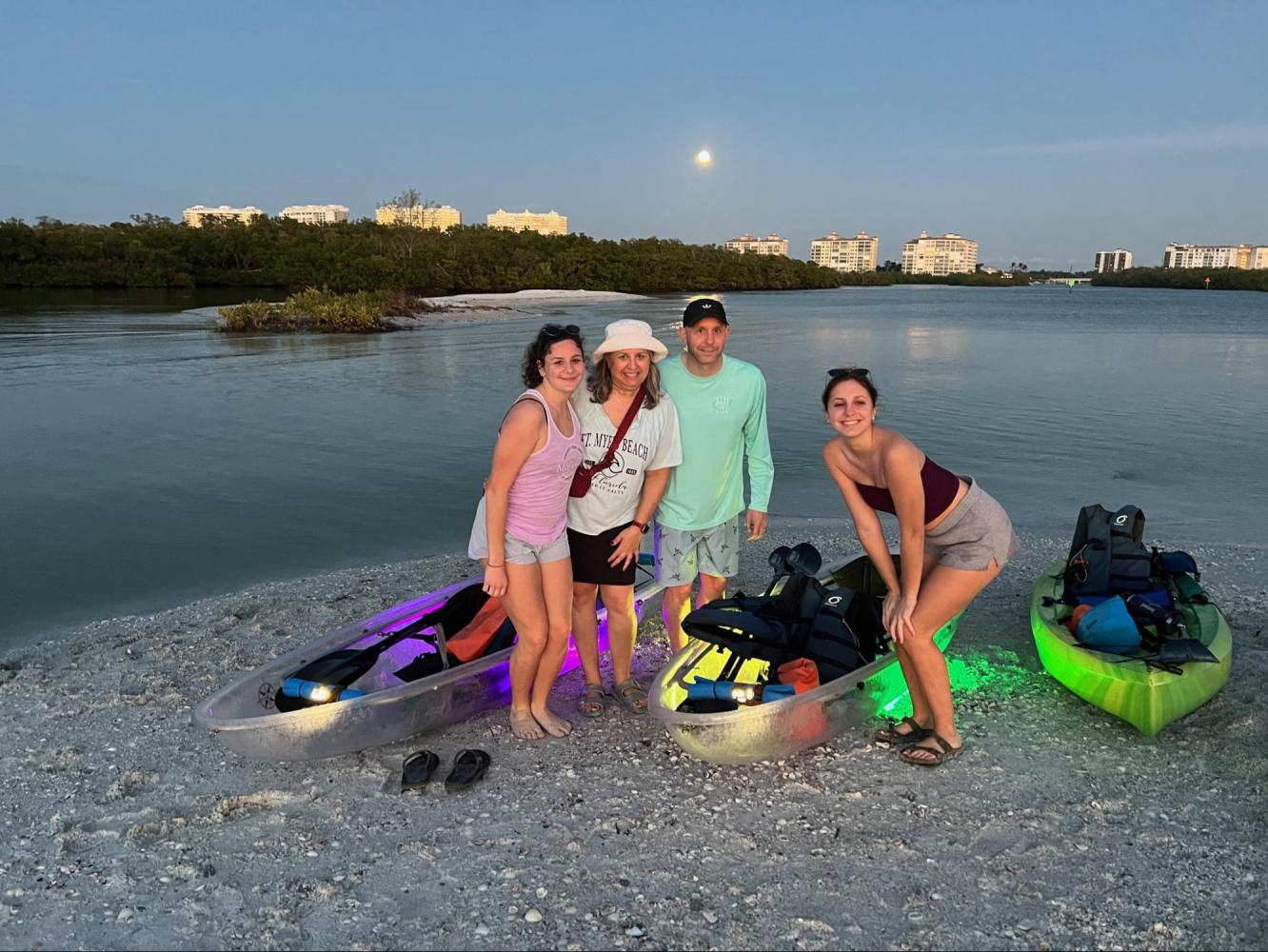 Four people stand by illuminated kayaks on a beach at dusk, with buildings and a full moon in the background.