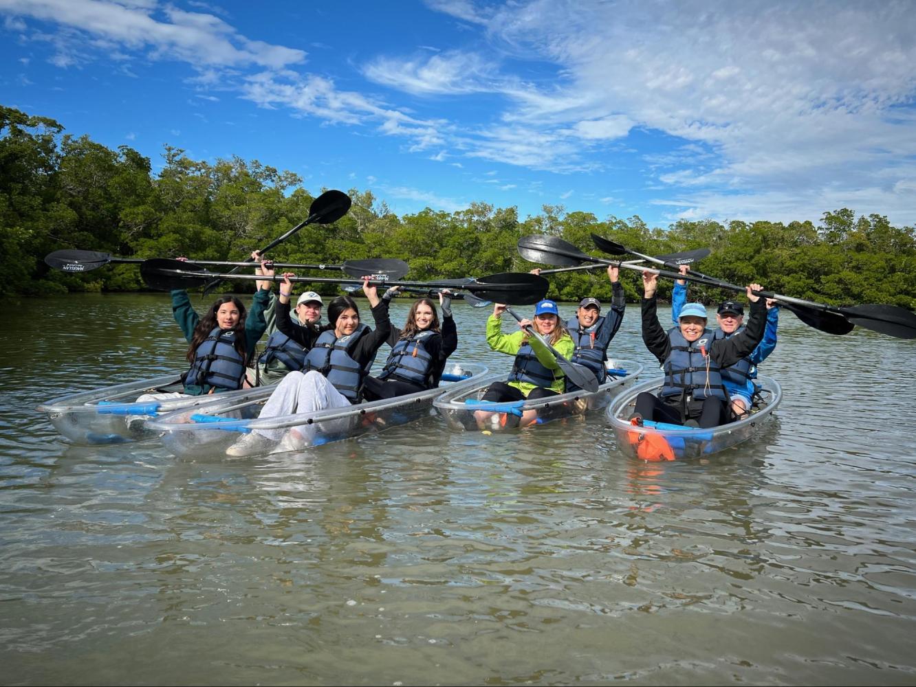 Group of people in clear kayaks lifting paddles on a calm river with blue sky and green trees.