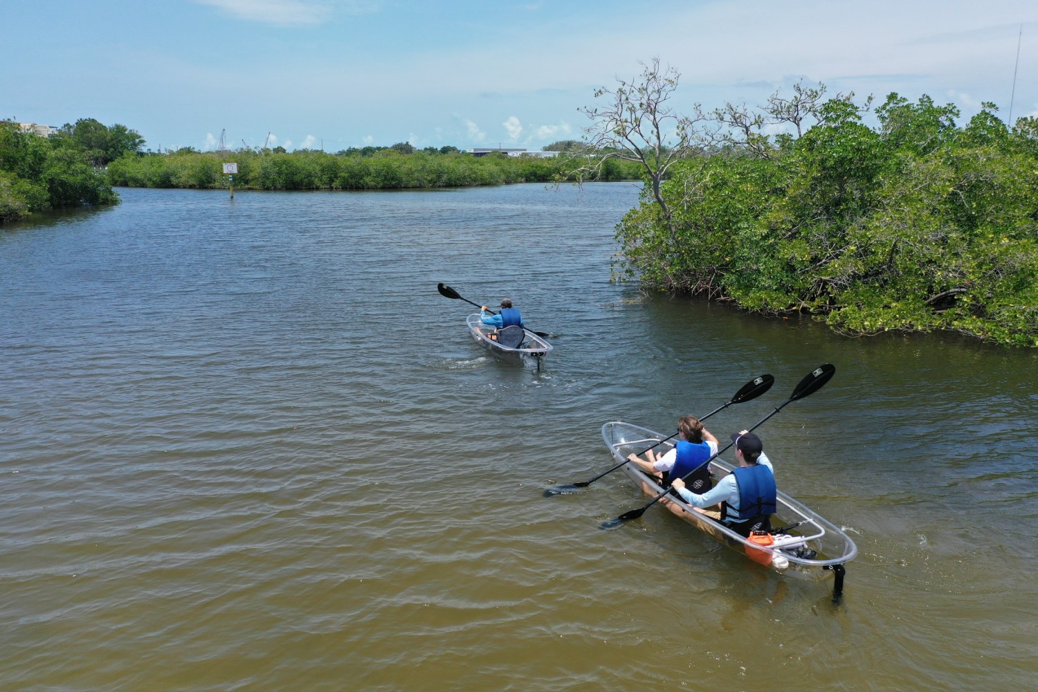 Two canoes with people paddling on a calm river surrounded by trees on a clear day.