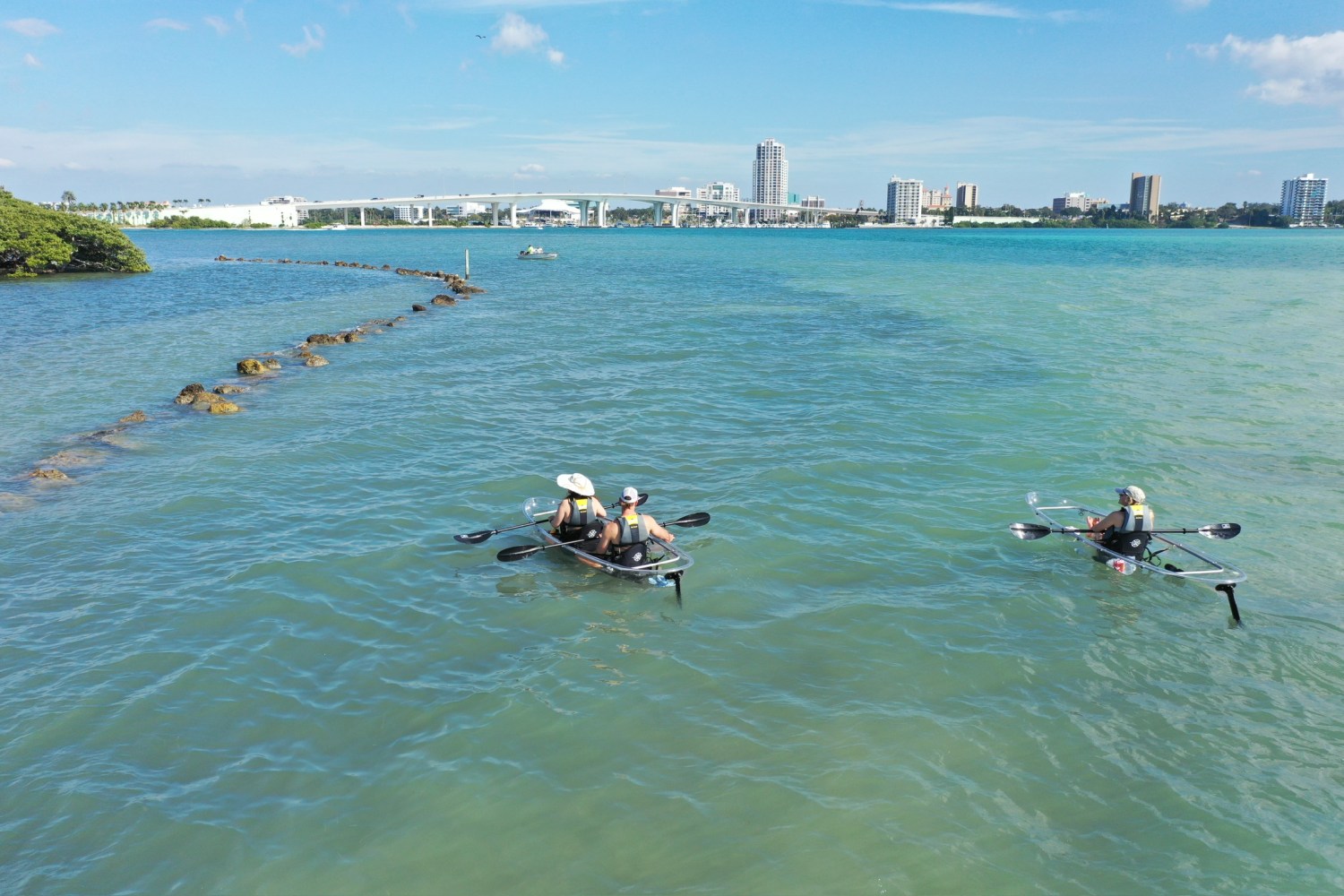 Two people kayaking on clear water near a city skyline under a blue sky.