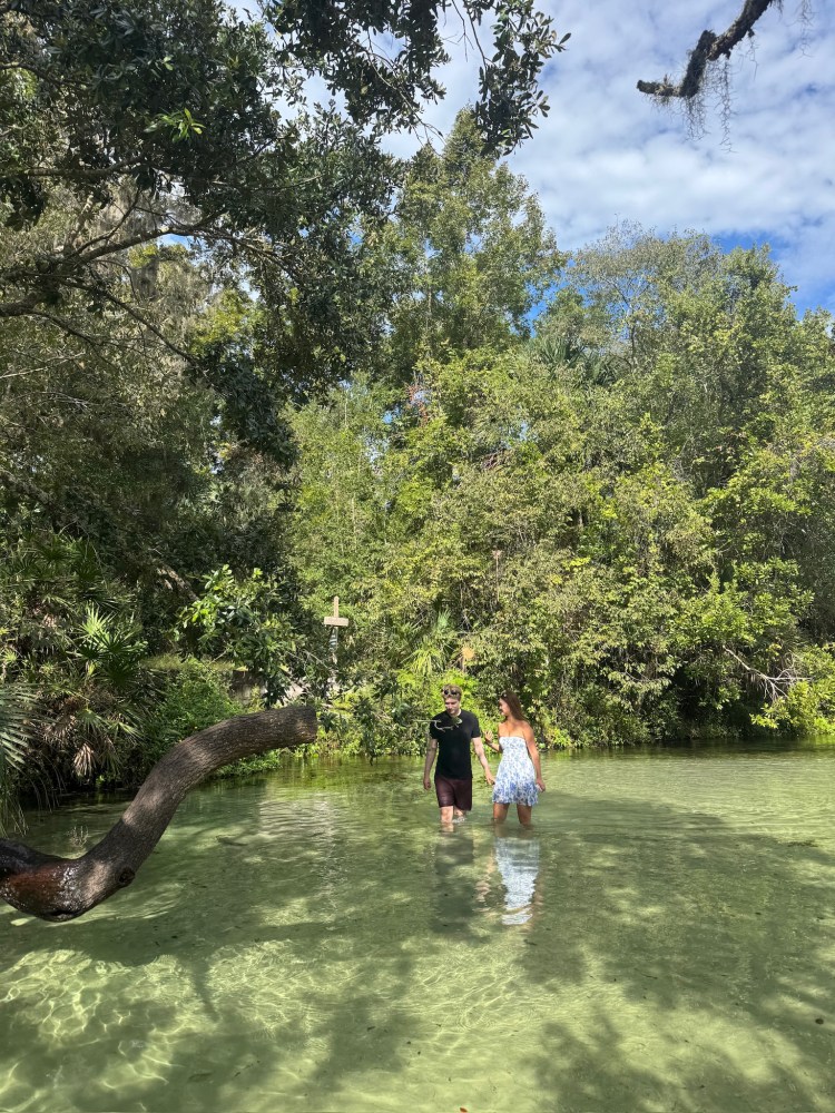 Two people wade in clear water surrounded by lush greenery and trees under a partly cloudy sky.