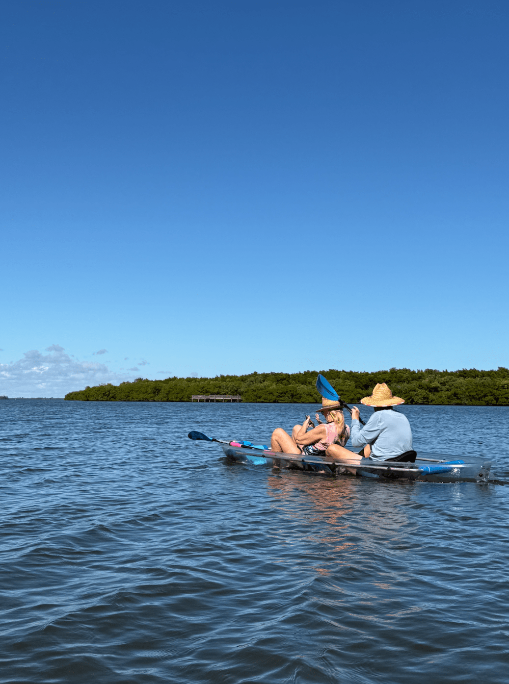 Two people kayaking on calm water under a clear blue sky, wearing hats and taking photos.