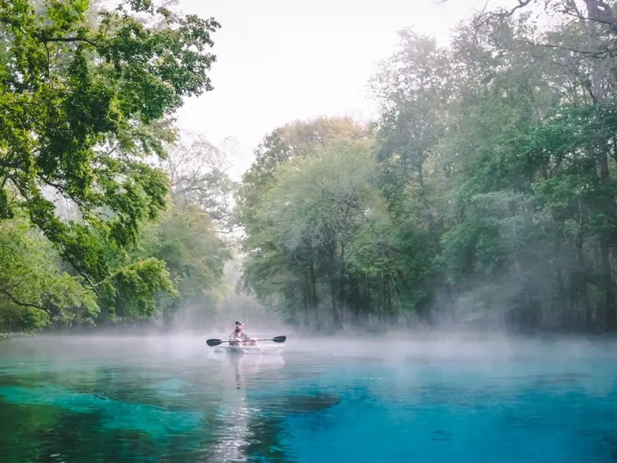 Person kayaking on a misty turquoise river surrounded by lush green trees.