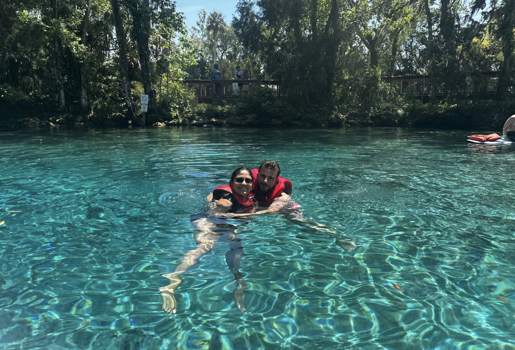 Two people in red life vests swimming in clear blue water surrounded by trees.