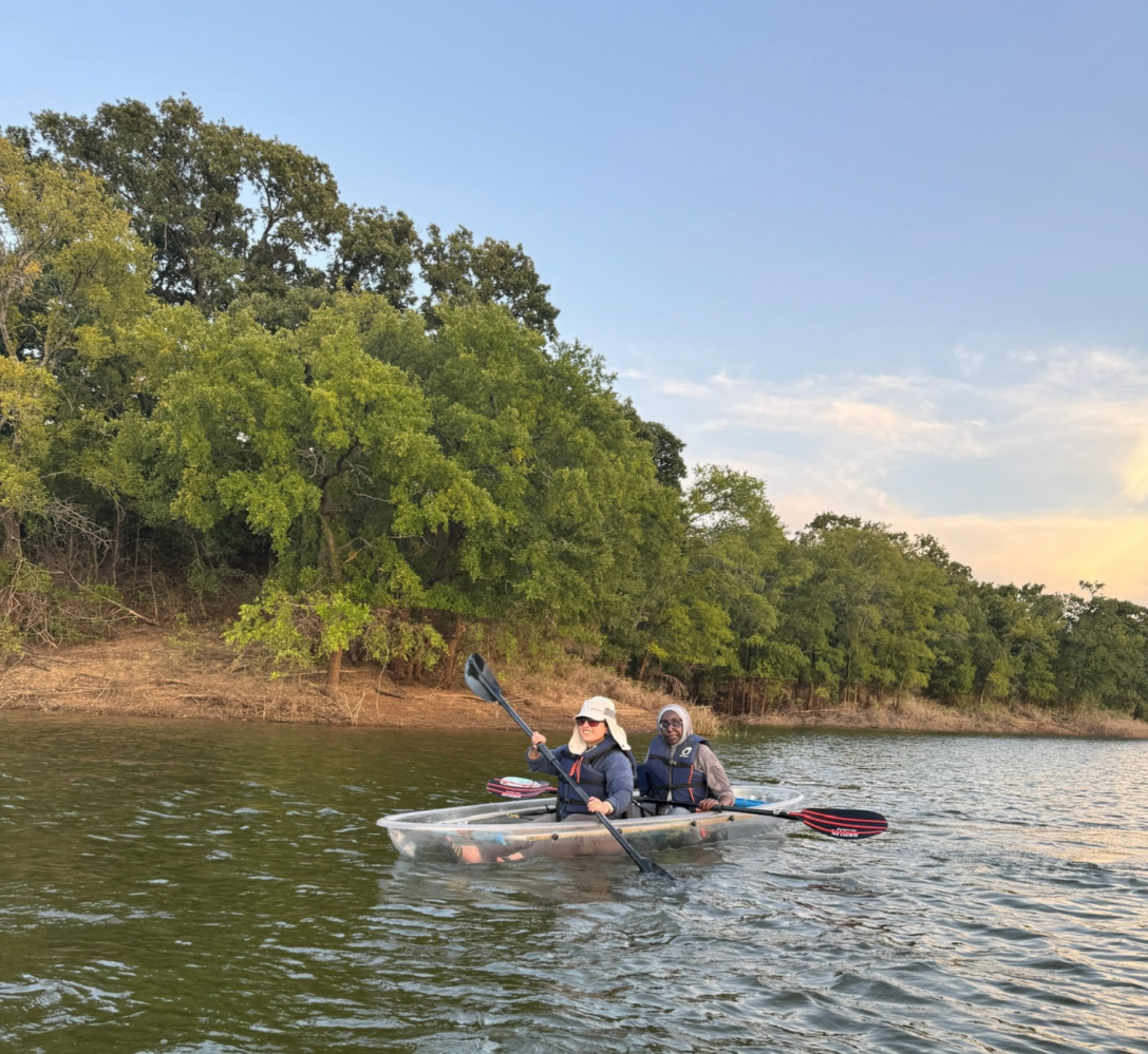 Two people kayaking on a lake near a tree-lined shore under a clear sky.