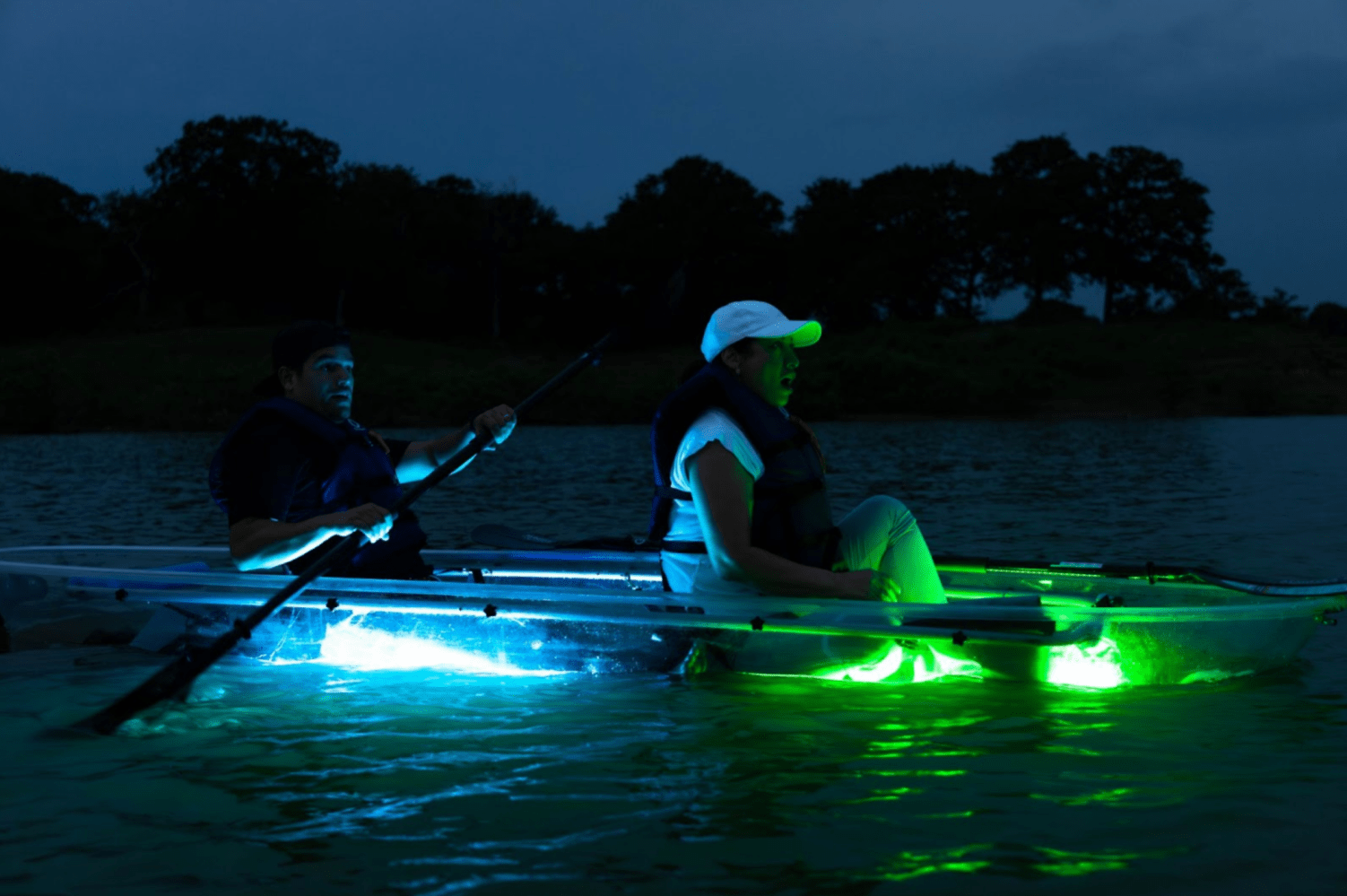 Two people in illuminated kayaks paddling on a lake at dusk.