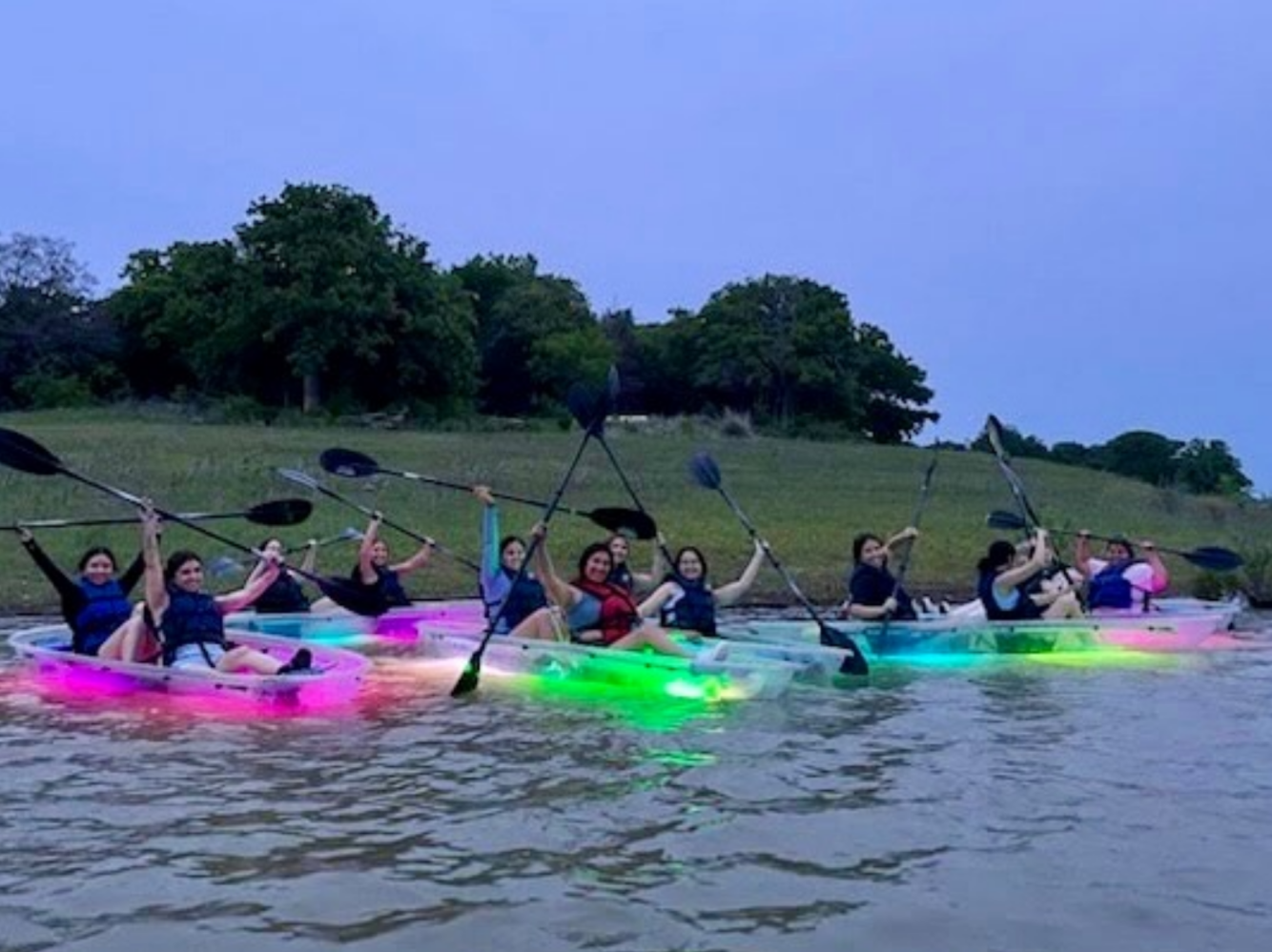 Group of people kayaking on lit-up kayaks with paddles raised, on a lake near a grassy shore at dusk.