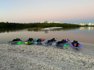 Six kayaks with colored lights on a pebble beach by calm water at sunset.