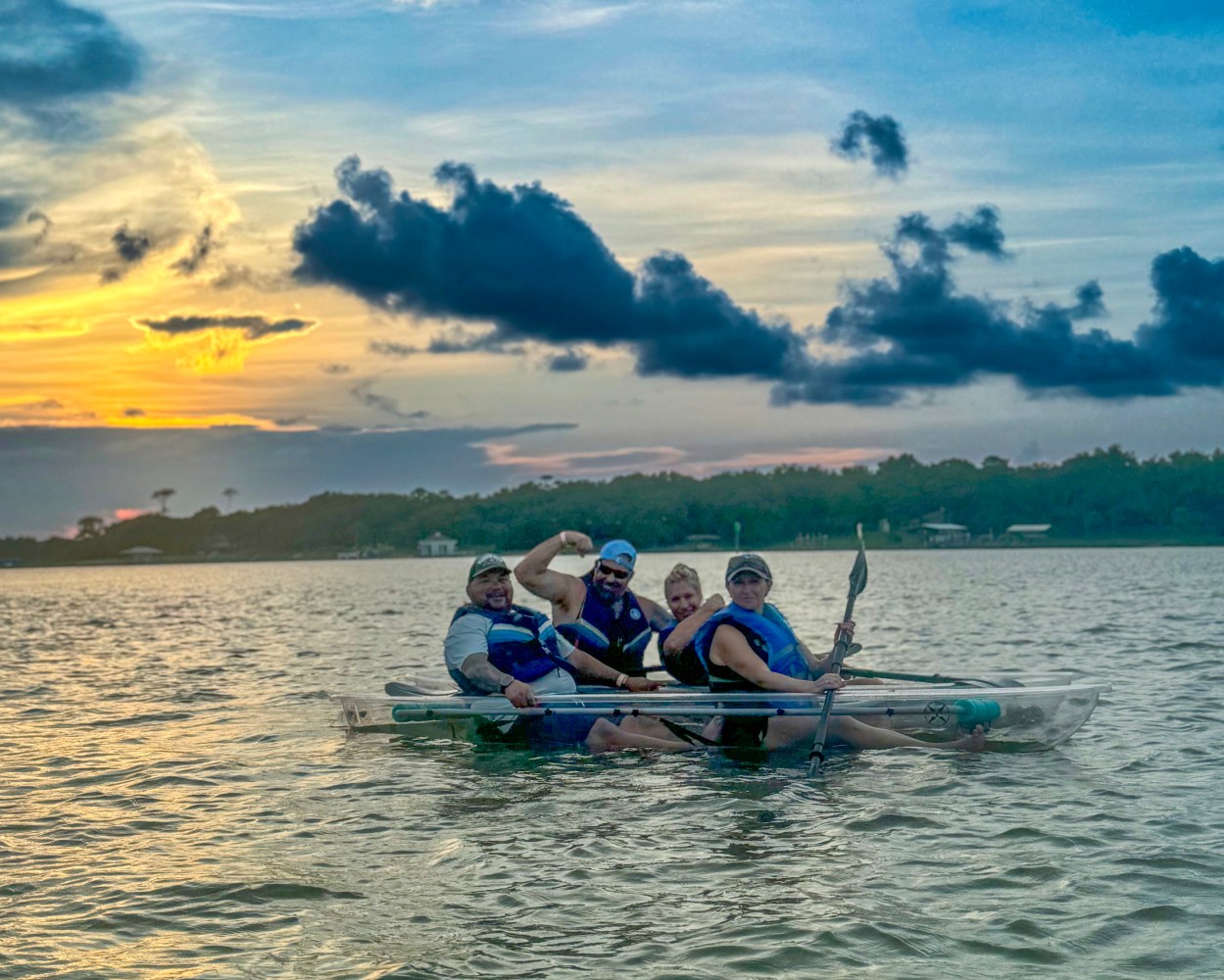 Four people in a transparent kayak on a lake at sunset.