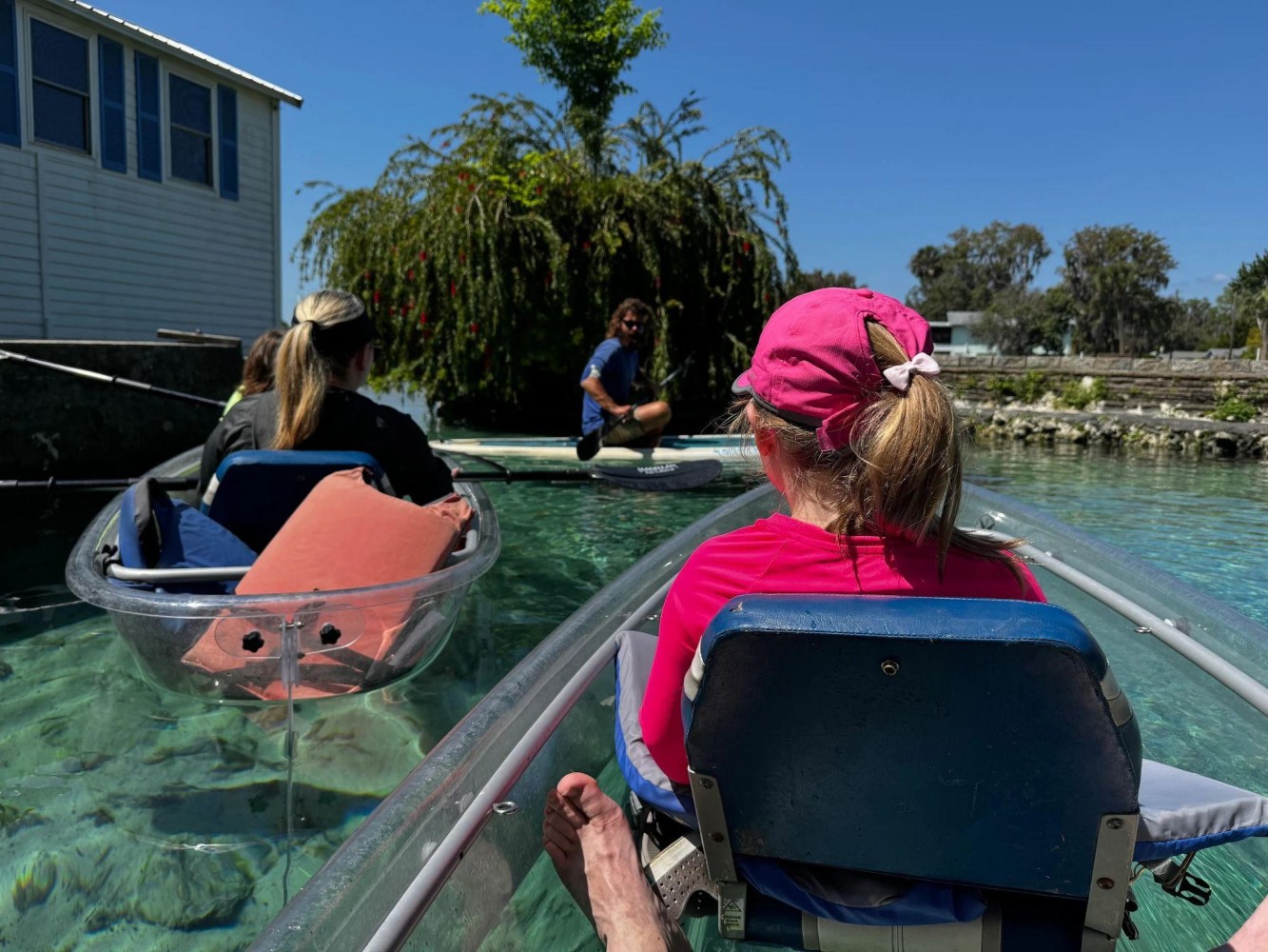 People in clear kayaks paddling near a house, with a person sitting on a dock.