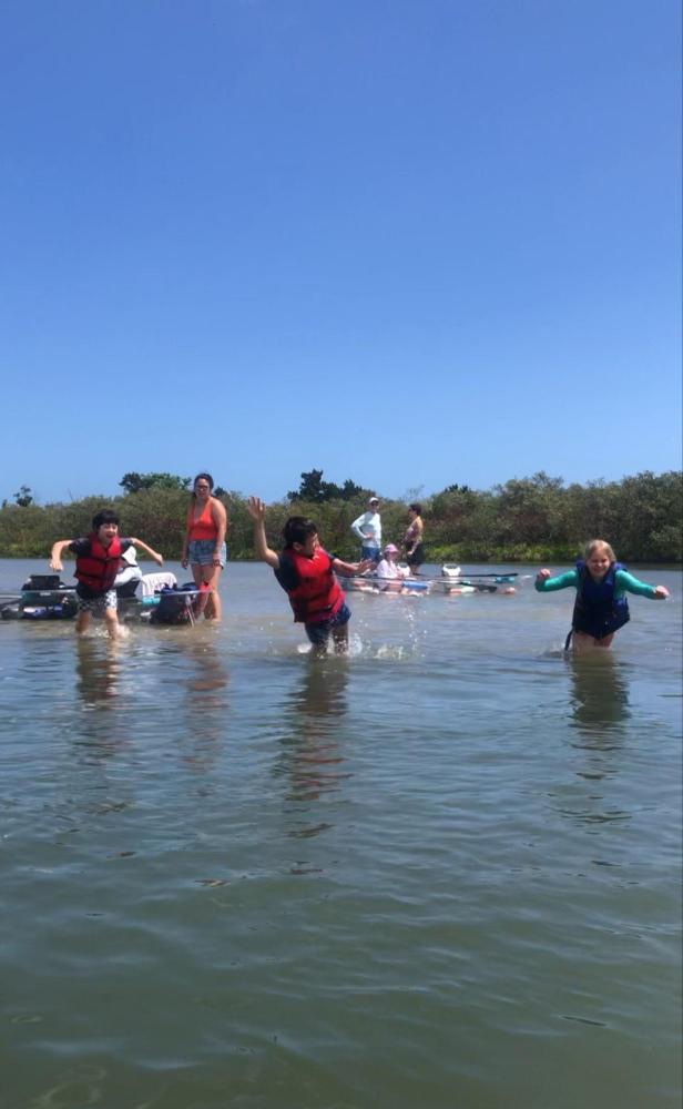 People in life vests playing in shallow water under a clear blue sky.