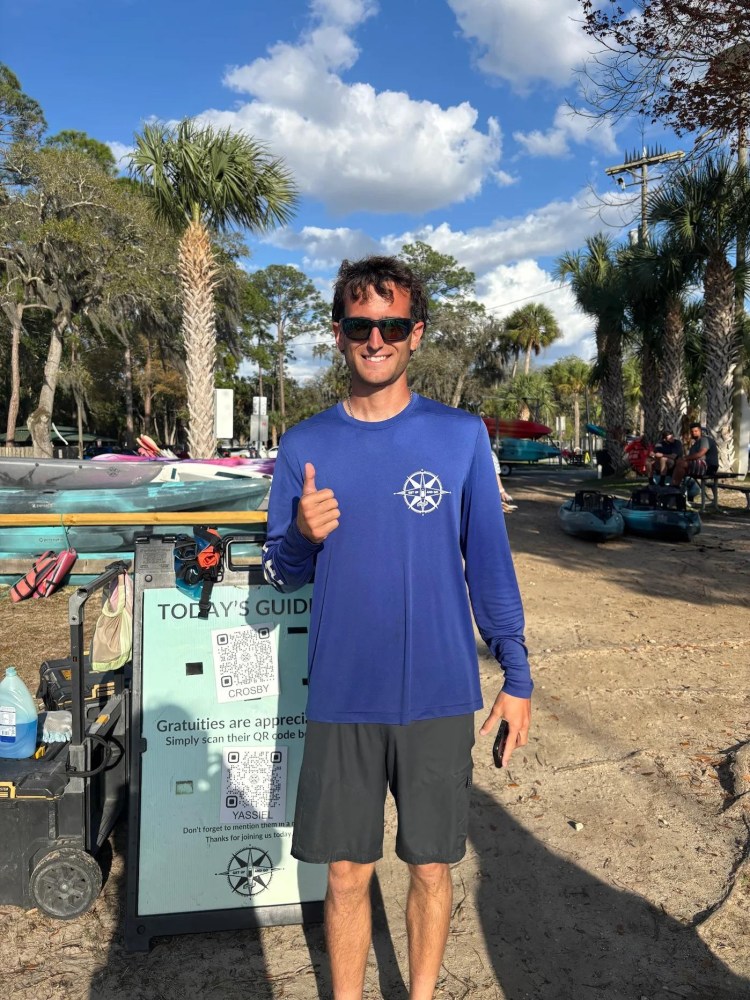 Man in blue shirt giving thumbs-up near a kayak rental booth with palm trees and blue sky in background.
