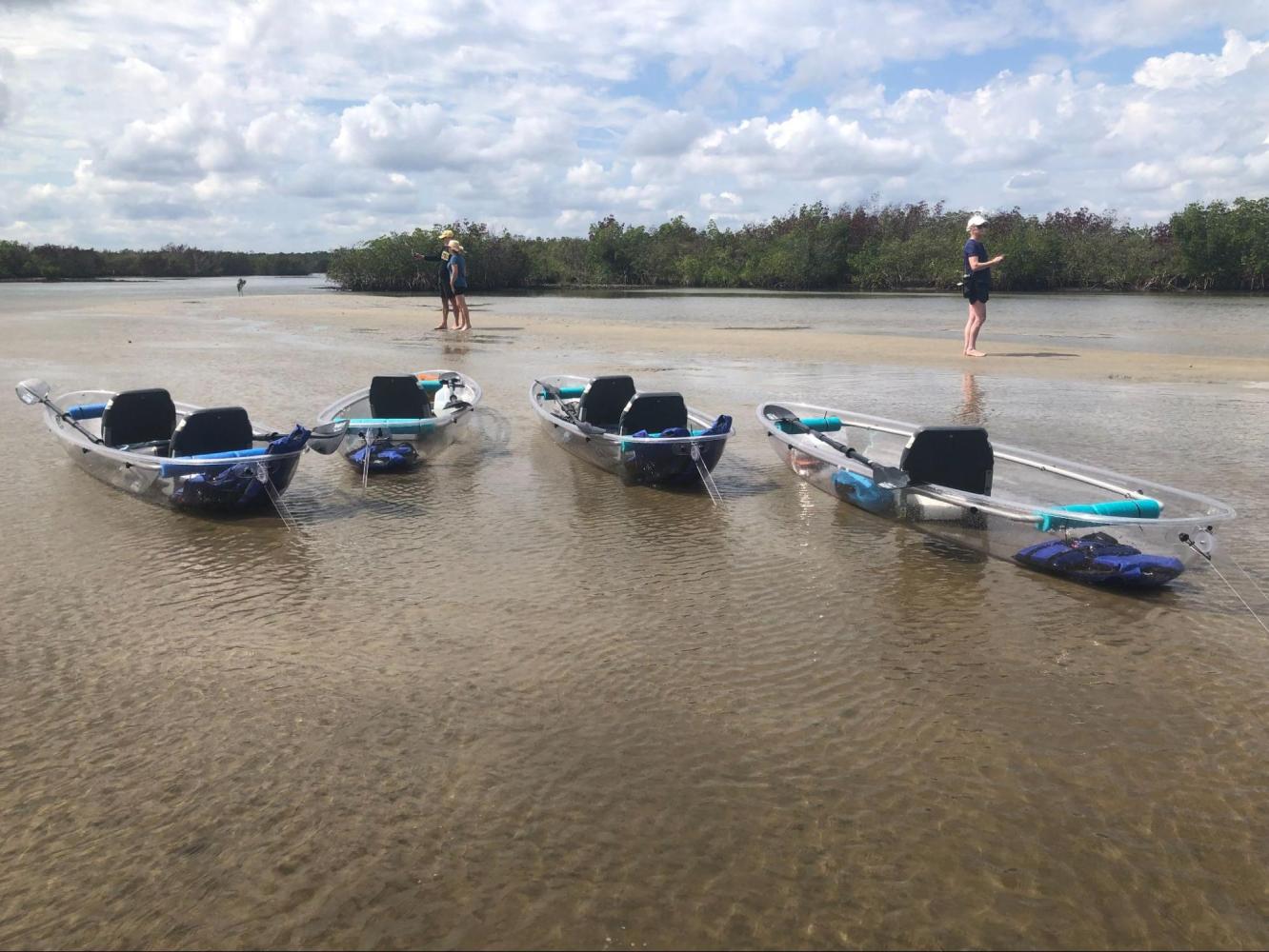 Four clear kayaks on shallow water with two people standing on a sandy shore.