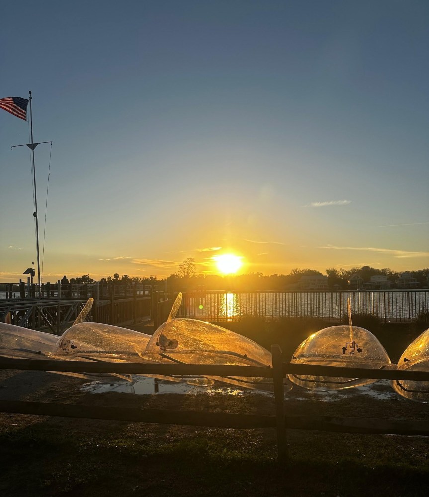 Transparent canoes by a lake at sunset with a flag and fence in the foreground.