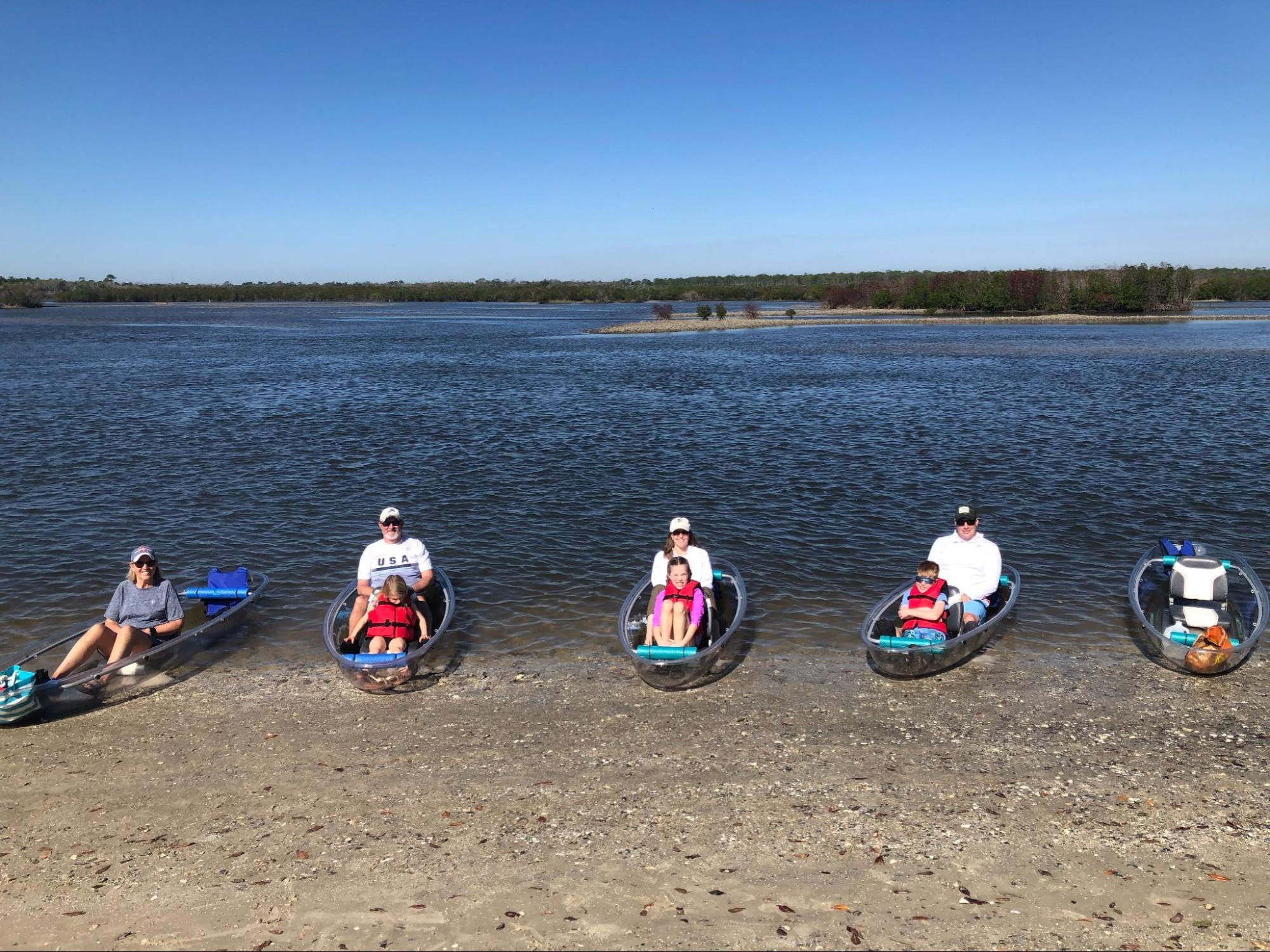 Six people in clear kayaks on a lake shore under a clear blue sky.