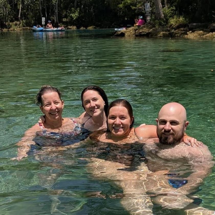 Four people smiling and standing in clear water, surrounded by greenery.
