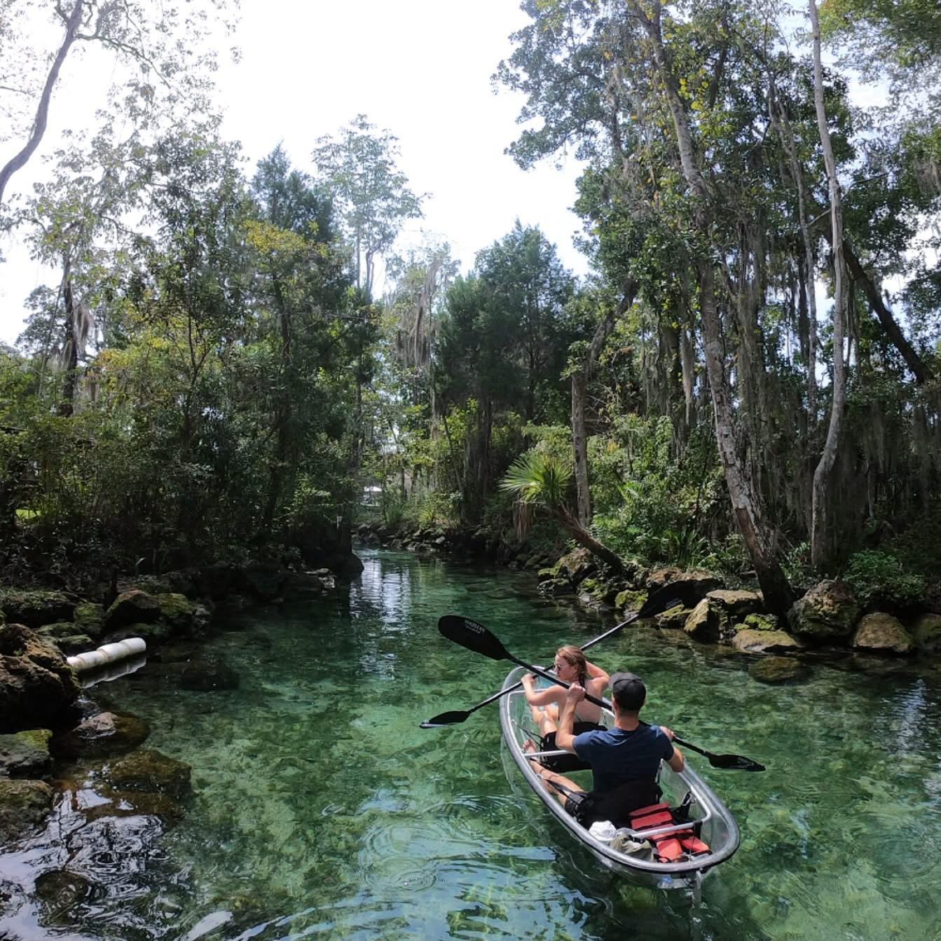 Two people kayaking in a clear, scenic river surrounded by lush greenery.