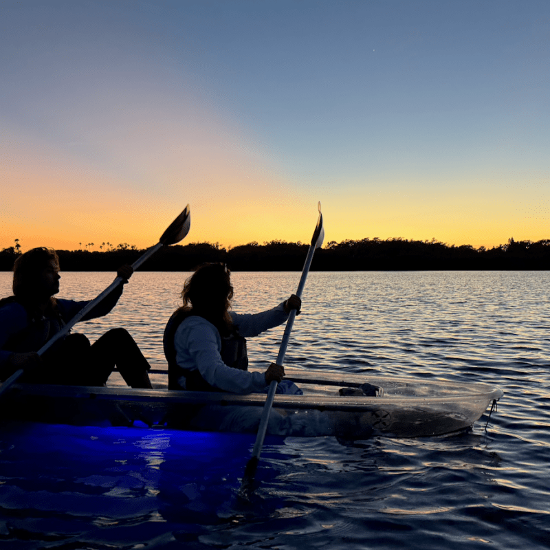 Two people kayaking on calm water at sunset, silhouetted against colorful sky.