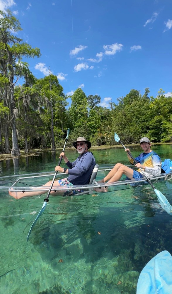 Two people kayaking in a clear kayak on a serene river.