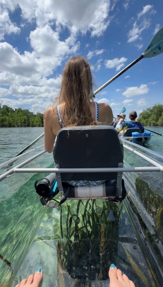 Woman on transparent kayak paddling on clear water, view from behind.