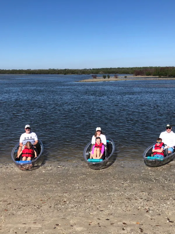 Five people sit in transparent kayaks by the shore on a sunny day.