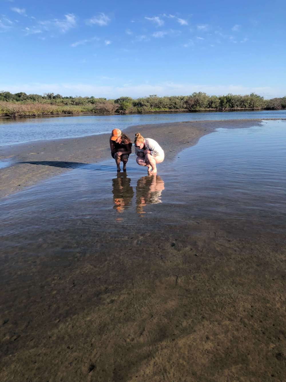 Two people squatting by shallow water on a sunny day.