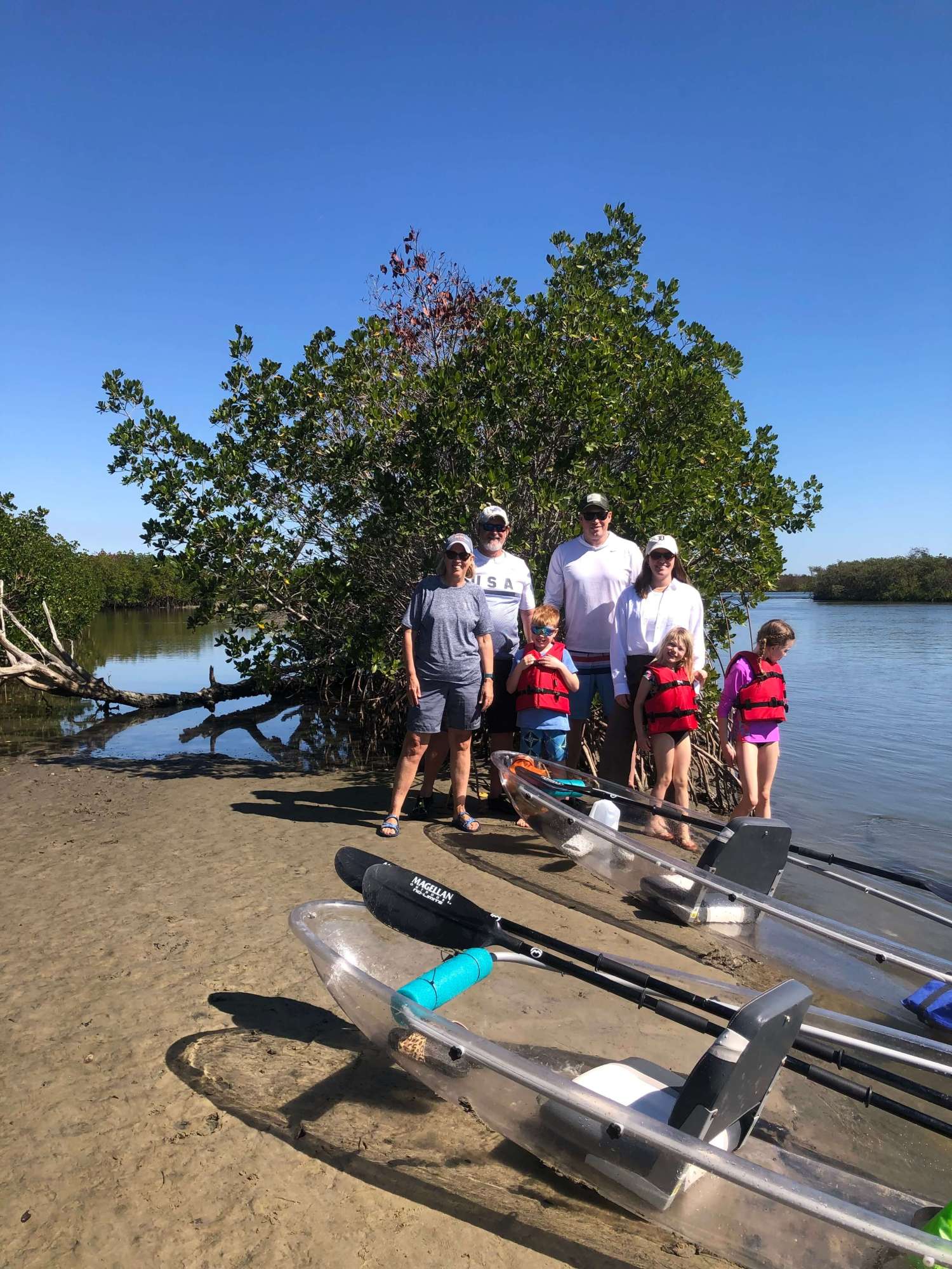 Family with clear kayaks by water, kids in life vests, trees in background, sunny day.