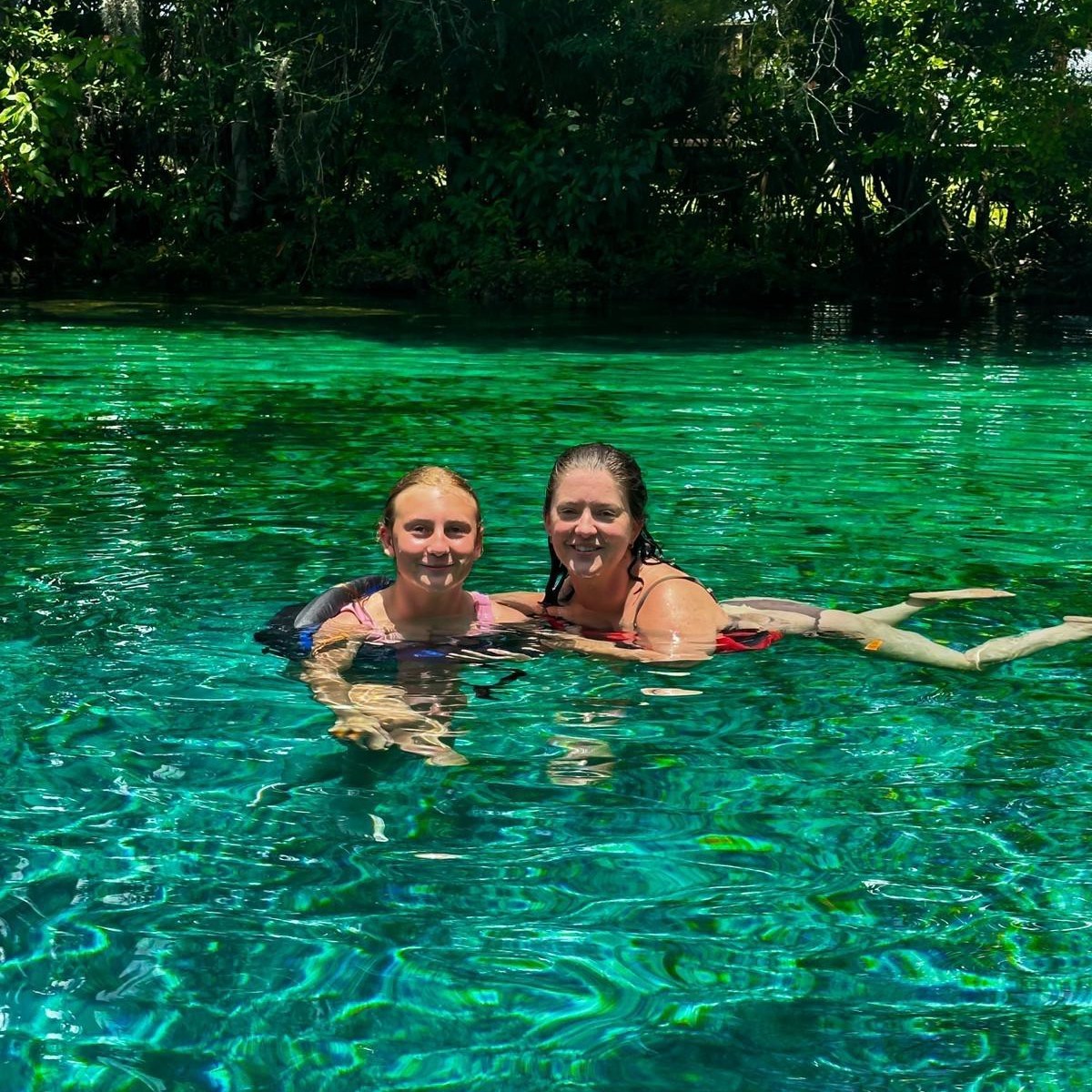 Two people swimming in clear turquoise water surrounded by trees.