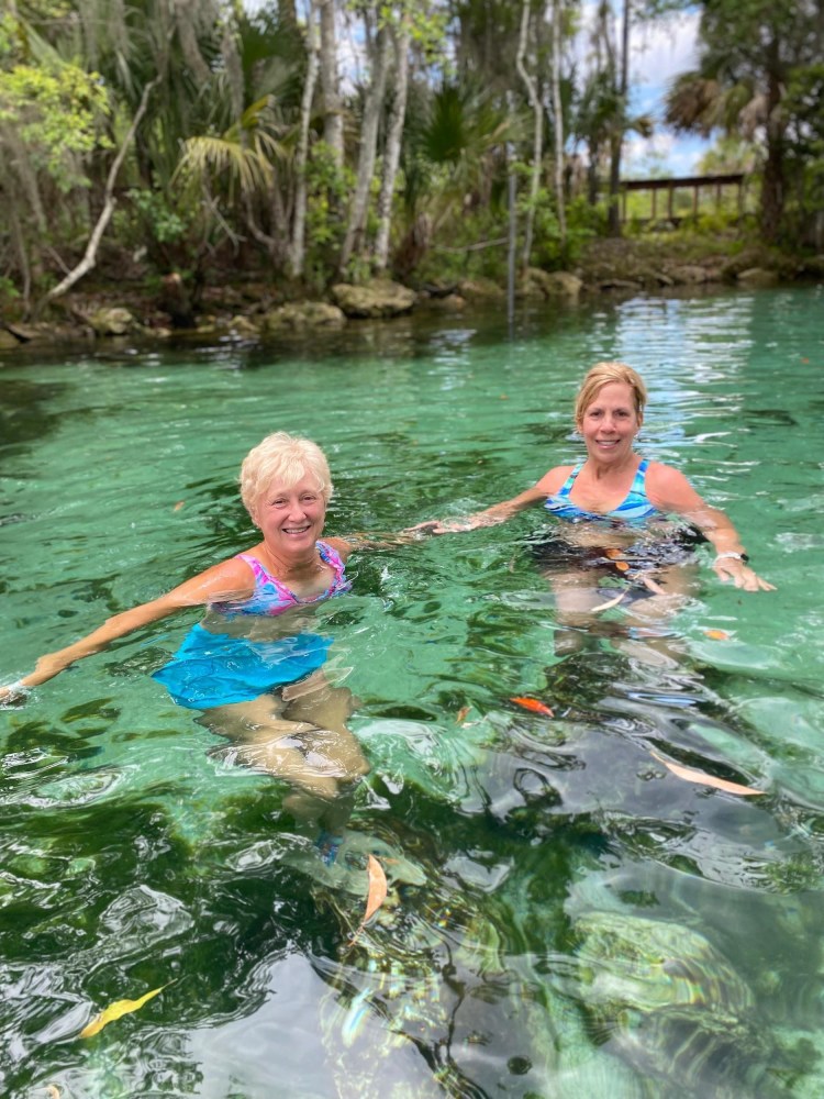 Two women swimming in a clear, green natural spring with surrounding trees.
