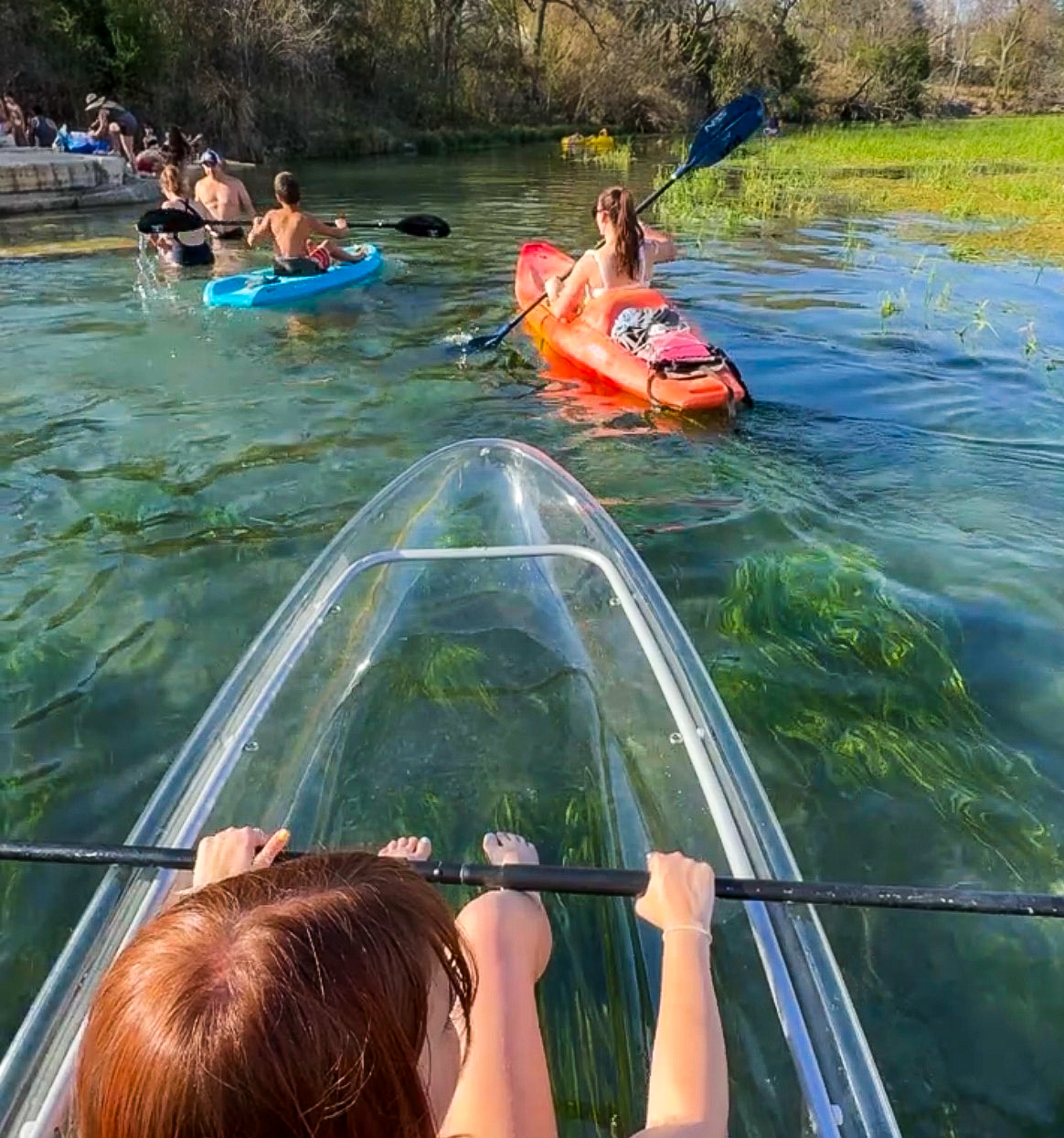 Person kayaking on clear water with others in the background, surrounded by greenery and clear skies.