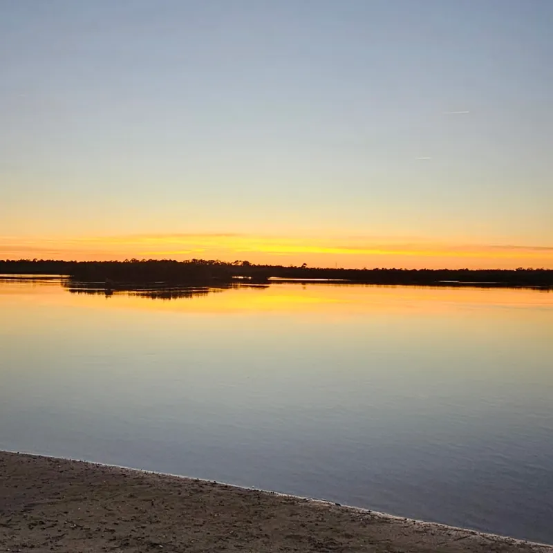 Calm lake at sunset with a sandy shore and orange sky.
