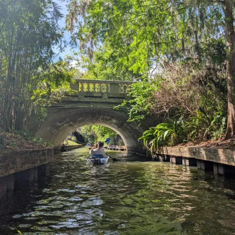 Person kayaking under a stone bridge on a lush, tree-lined waterway.