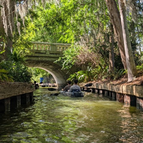 People kayaking under a stone bridge in a lush, narrow waterway.