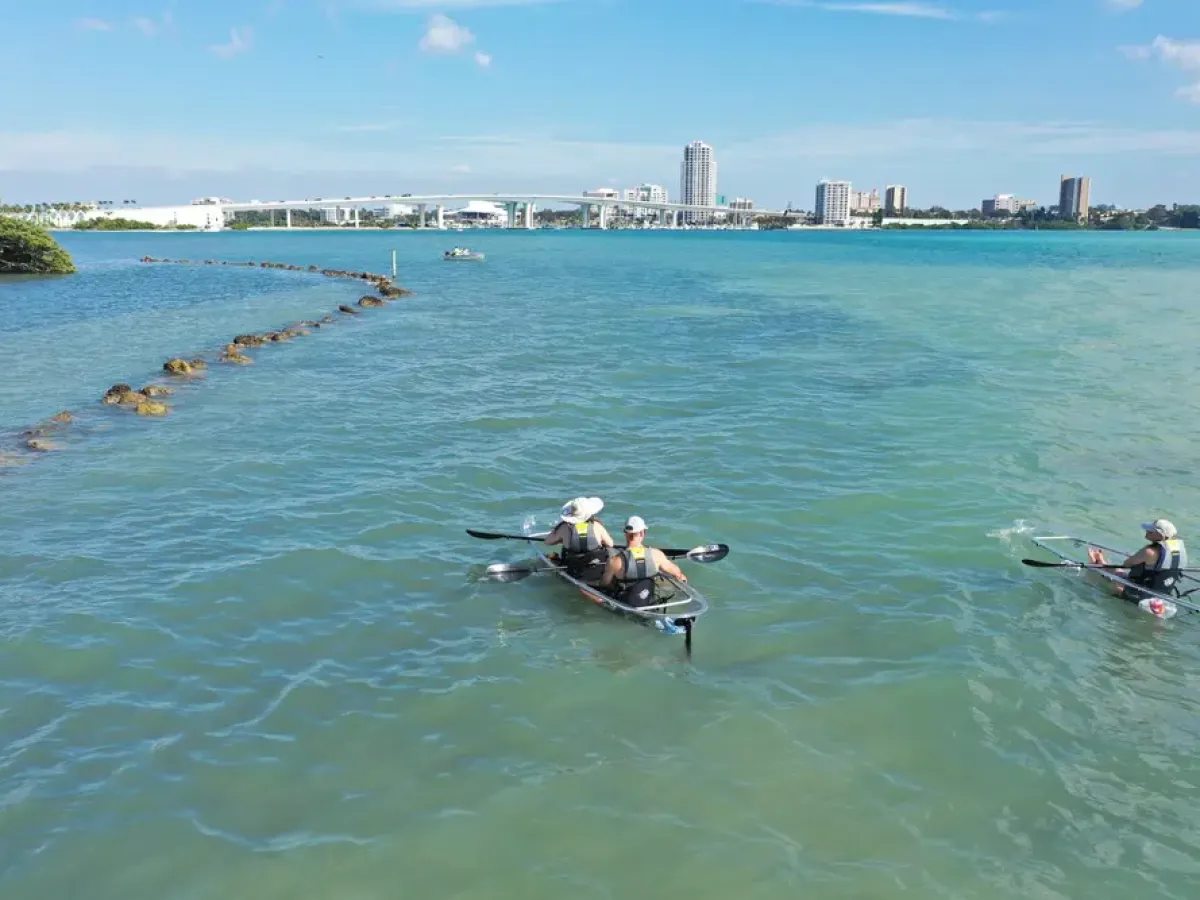 Two kayakers paddle in clear blue water near a city skyline.