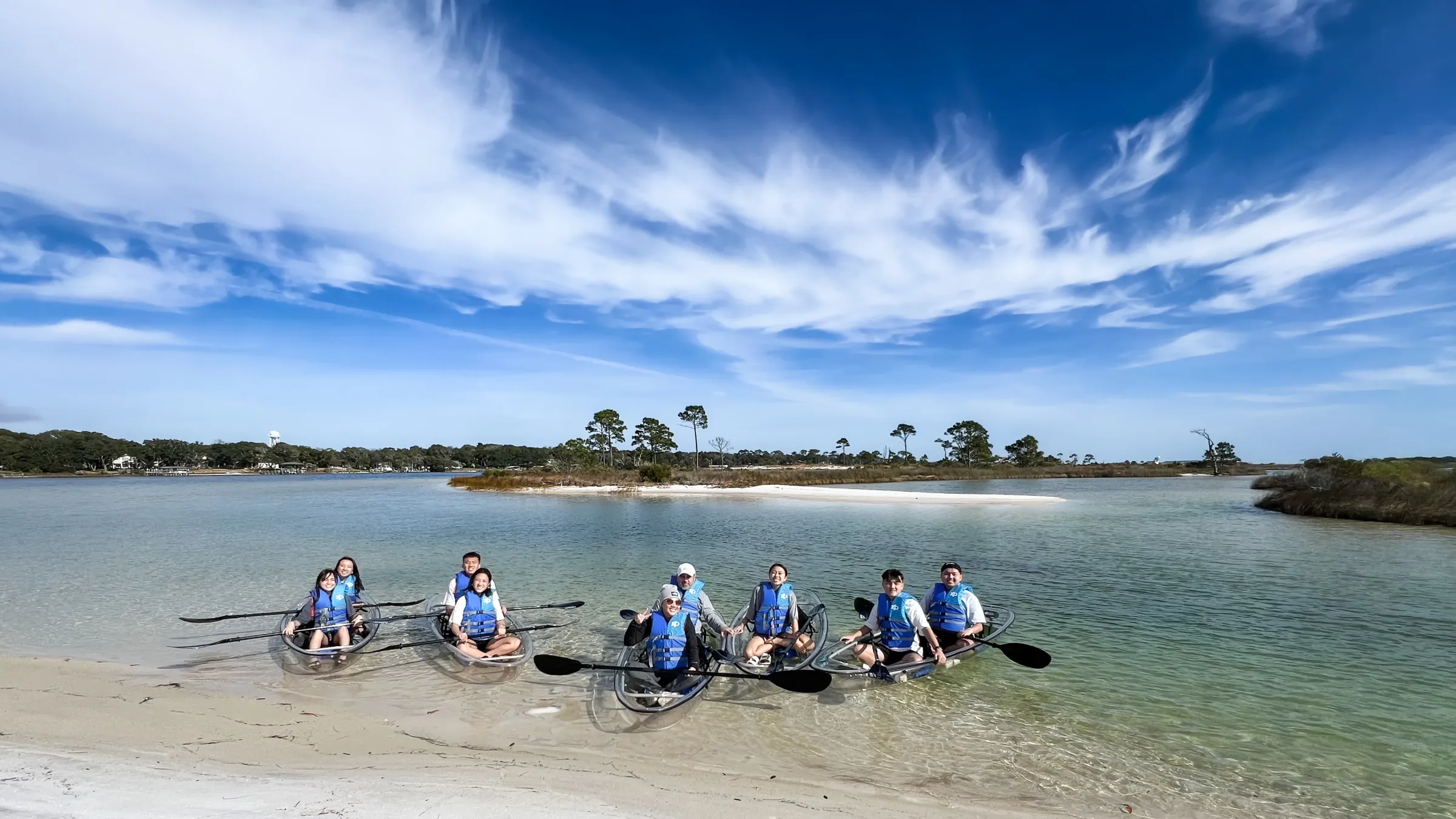 Group kayaking in clear boats on a sunny day, sandy shore, blue skies.