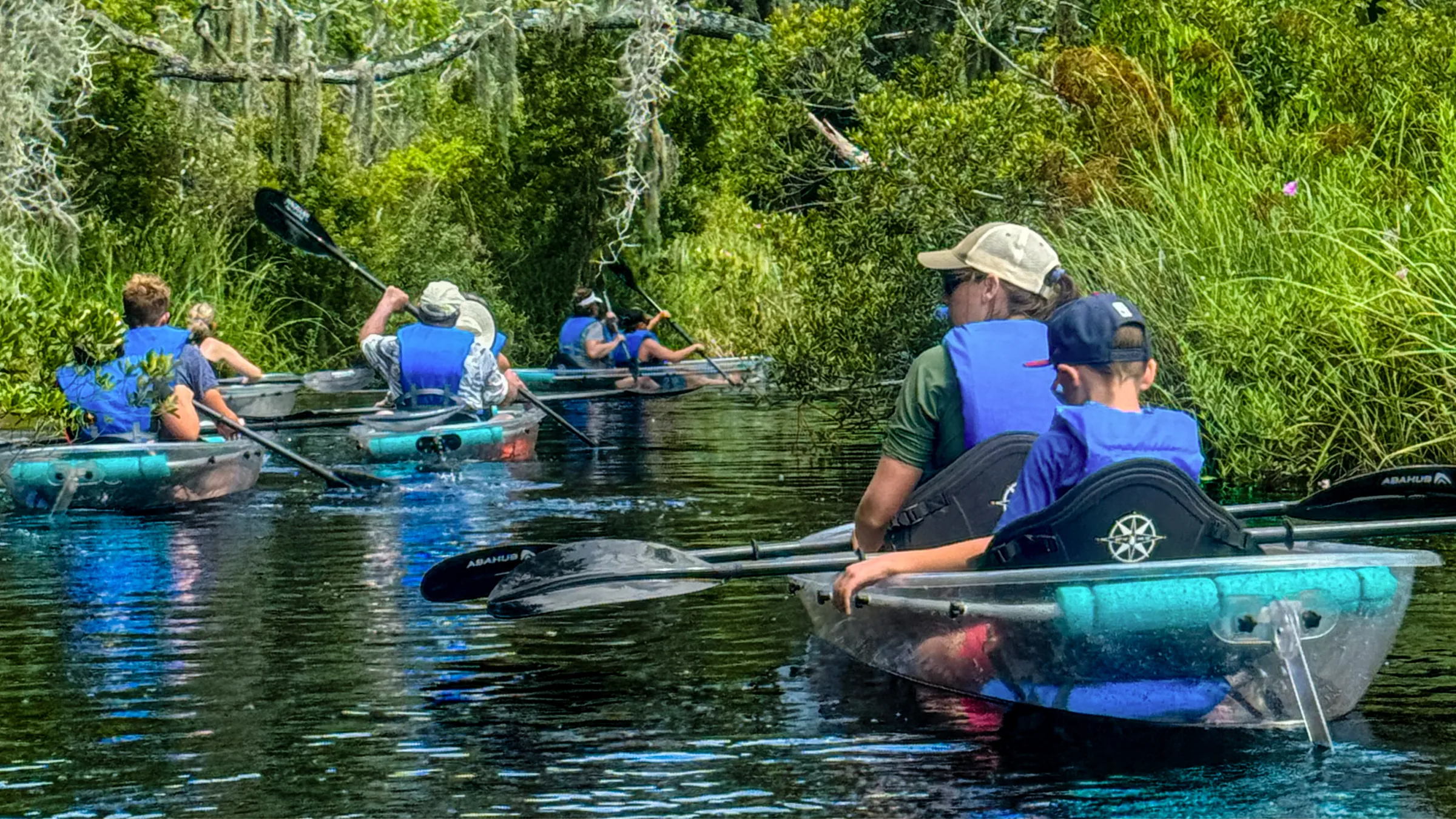 Group kayaking on a narrow forested river wearing blue life jackets.