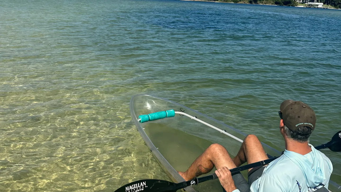 Man in clear kayak on calm, shallow water with distant shoreline and blue sky.