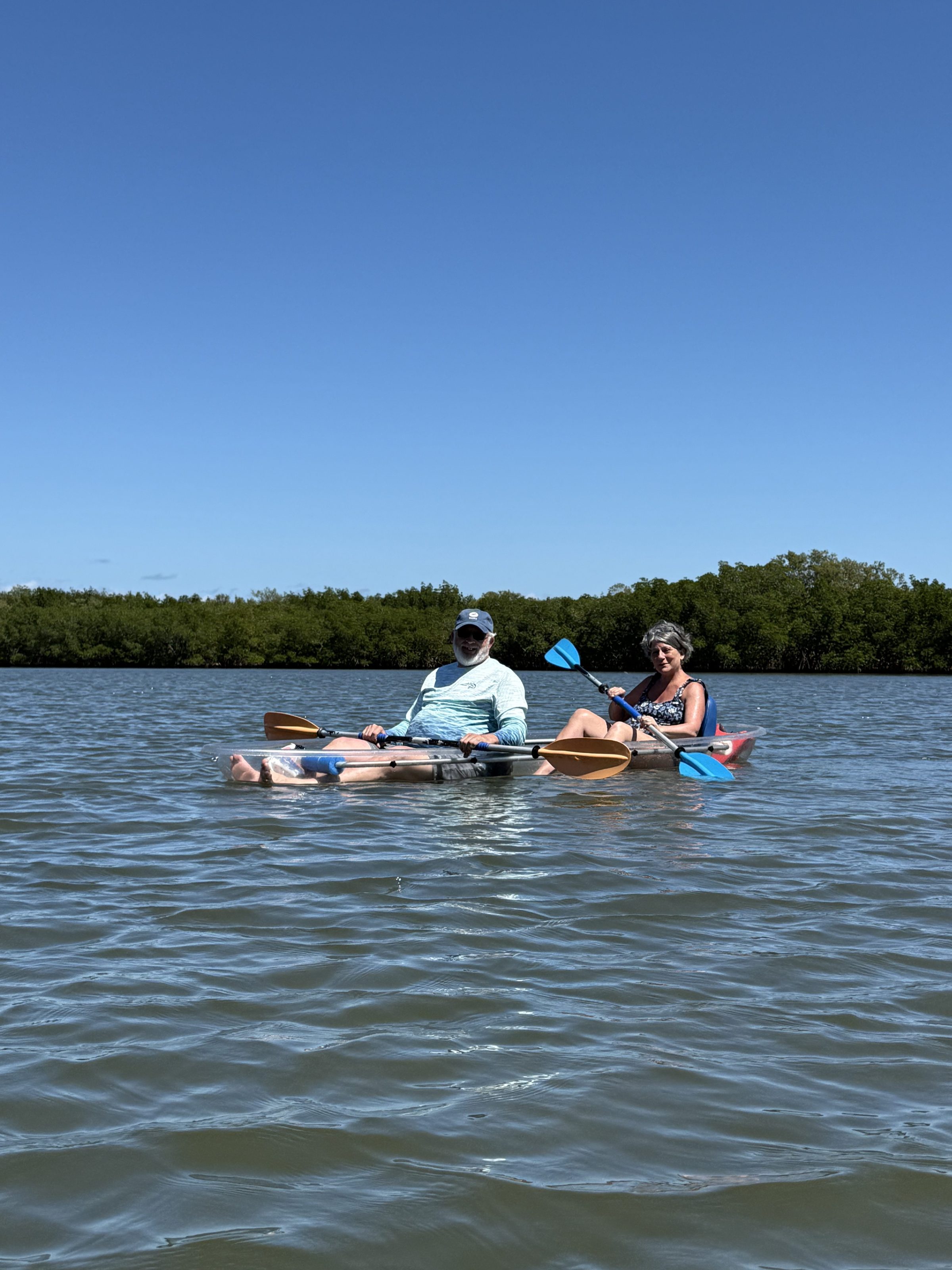 Two people kayaking on a calm lake with trees in the background under a clear blue sky.