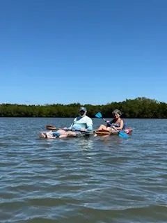 Two people kayaking on calm water near a forested shore.