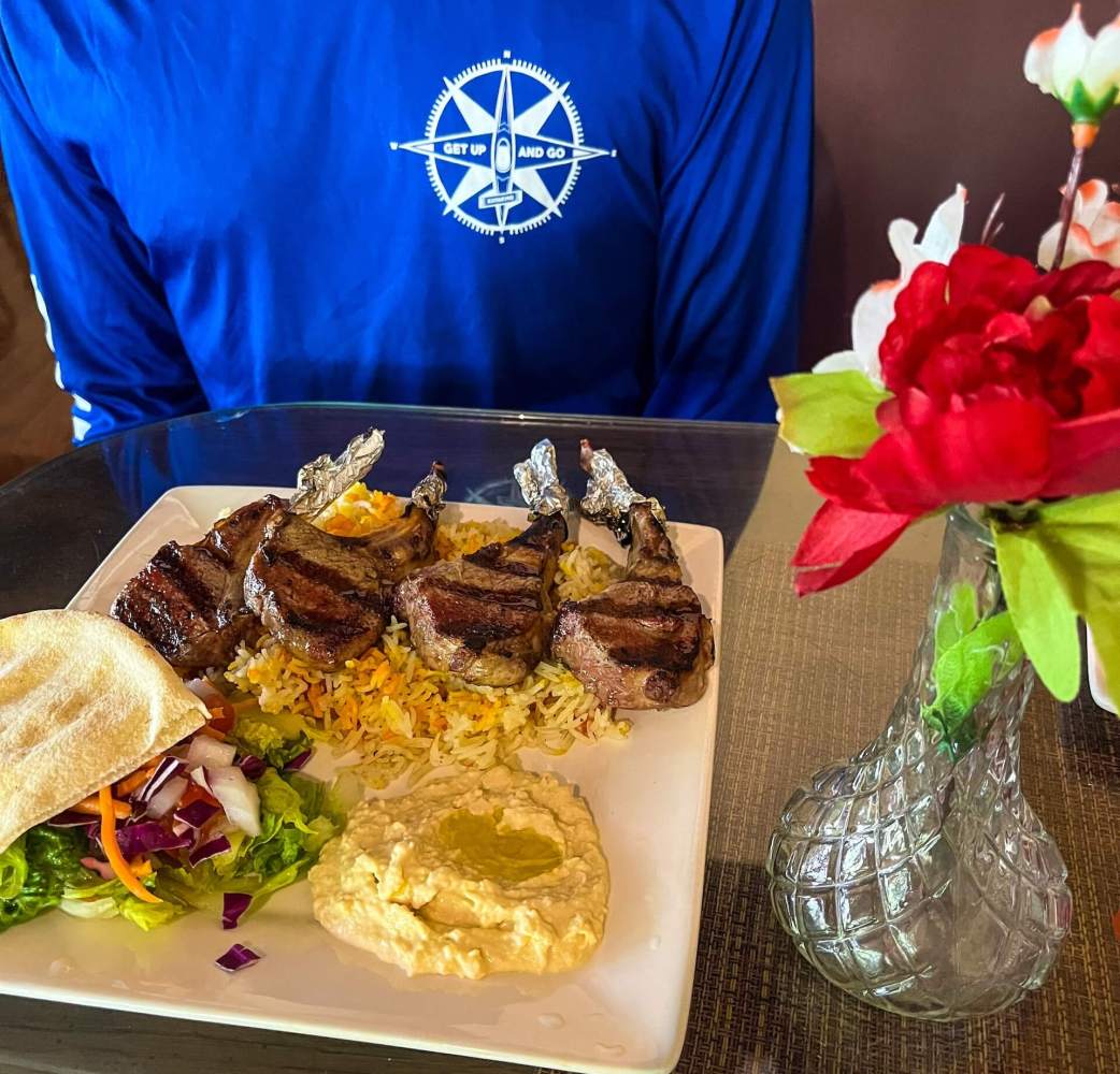 Plate with grilled lamb chops, salad, and hummus on a table beside a vase with red flowers.