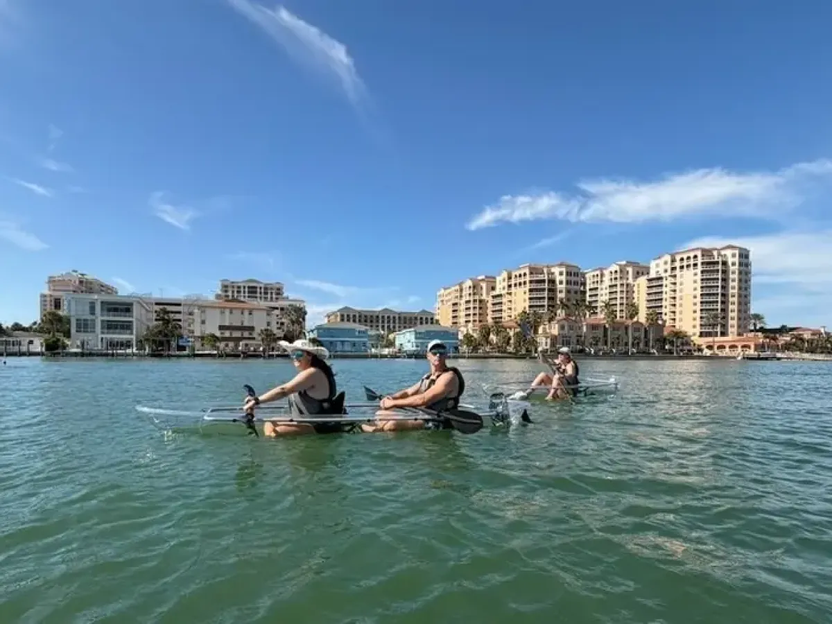 Two people kayaking on a calm sea with city buildings in the background.