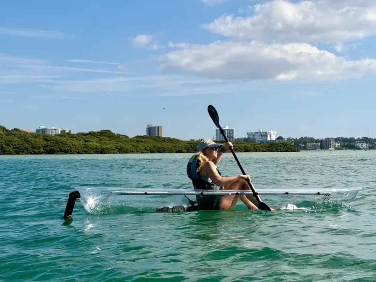 Person kayaking on clear kayak in calm waters, city skyline and trees in background.