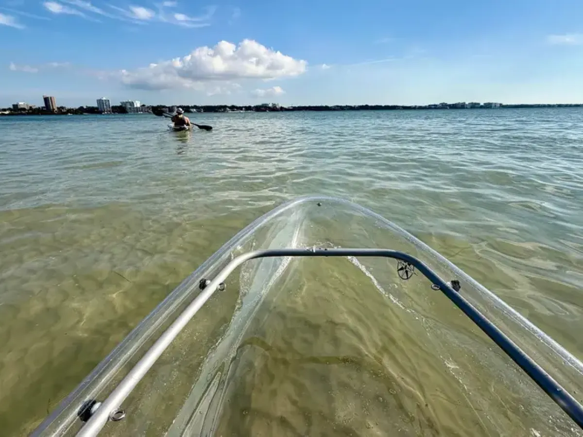 View from transparent kayak on calm sea towards city skyline, under a partly cloudy sky.