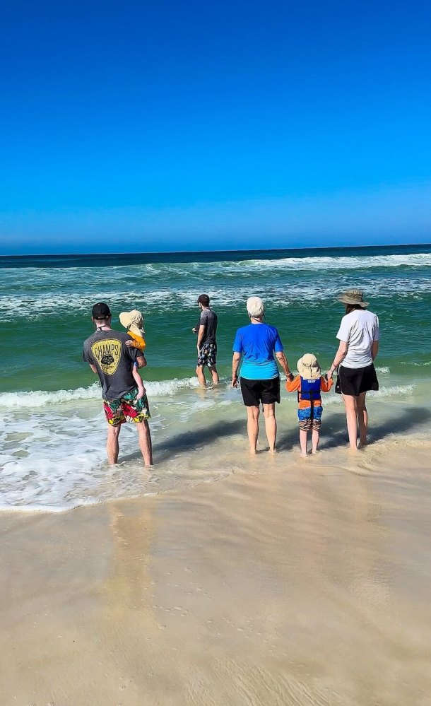 People on a sandy beach, some wading into the ocean under a clear blue sky.