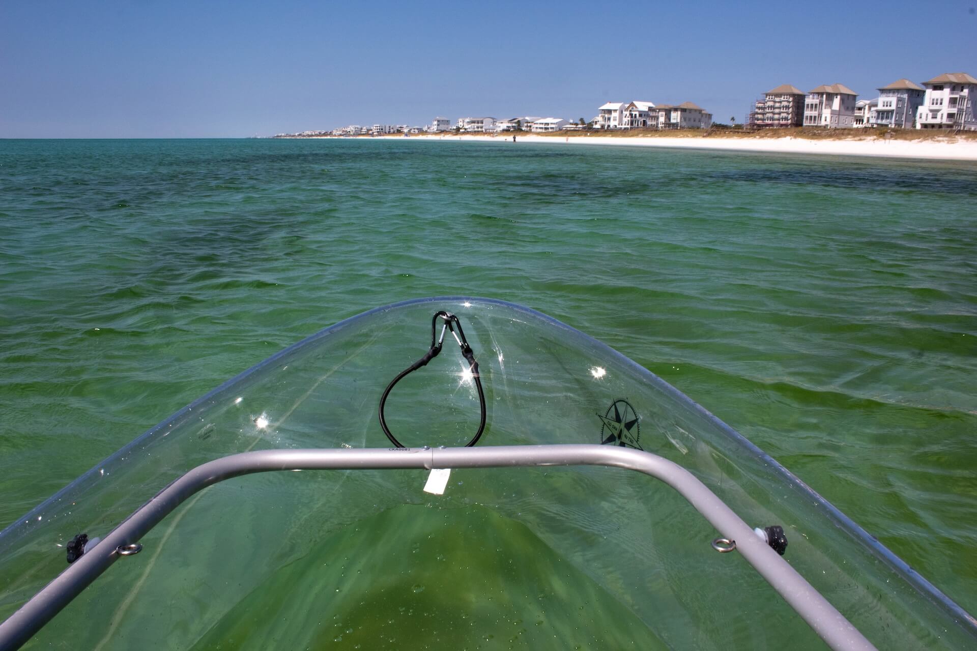 View from clear kayak on green water towards beach with houses.