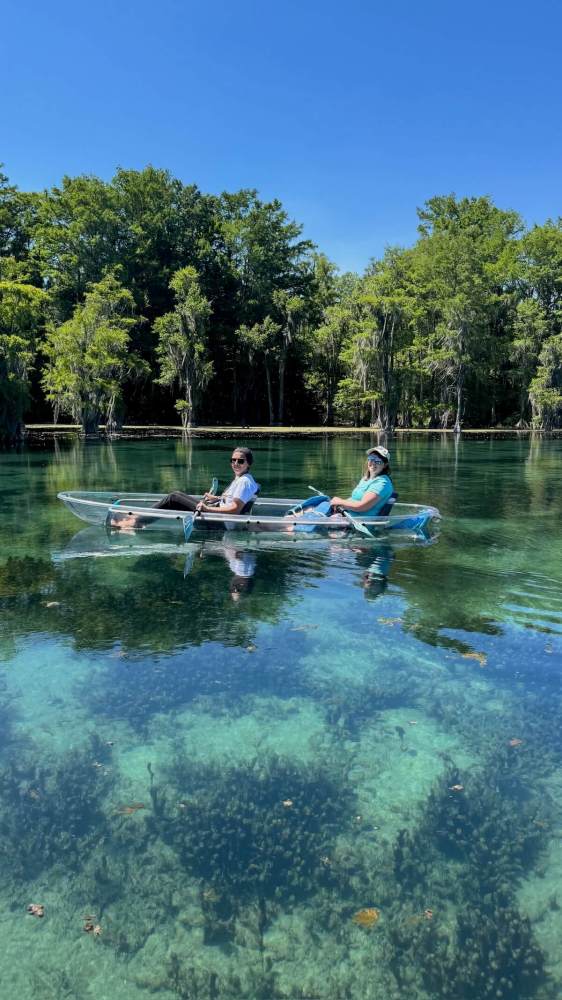 Two people in a clear kayak on a calm, clear lake with trees in the background.