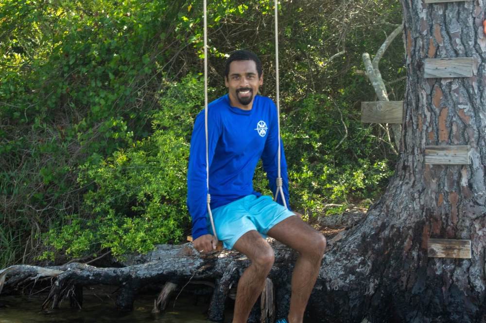 Man in blue outfit sitting on a tree swing by water and greenery.