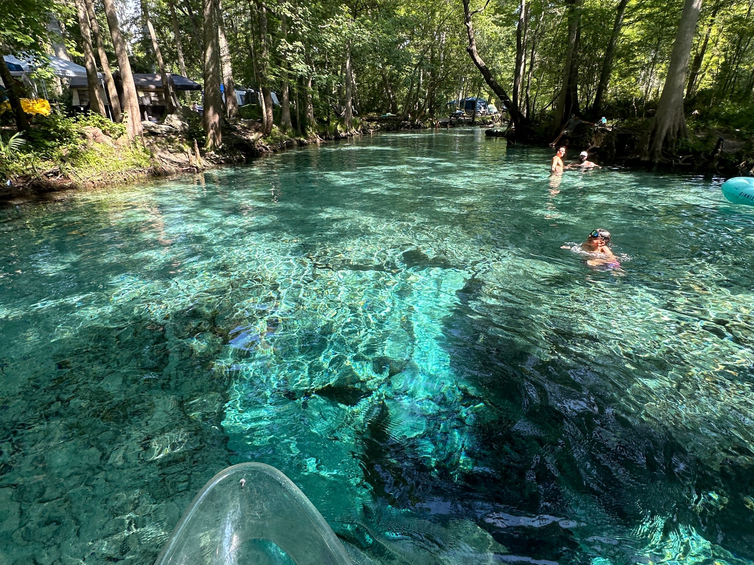 Clear kayak on a crystal-clear river with swimmers and lush green trees.