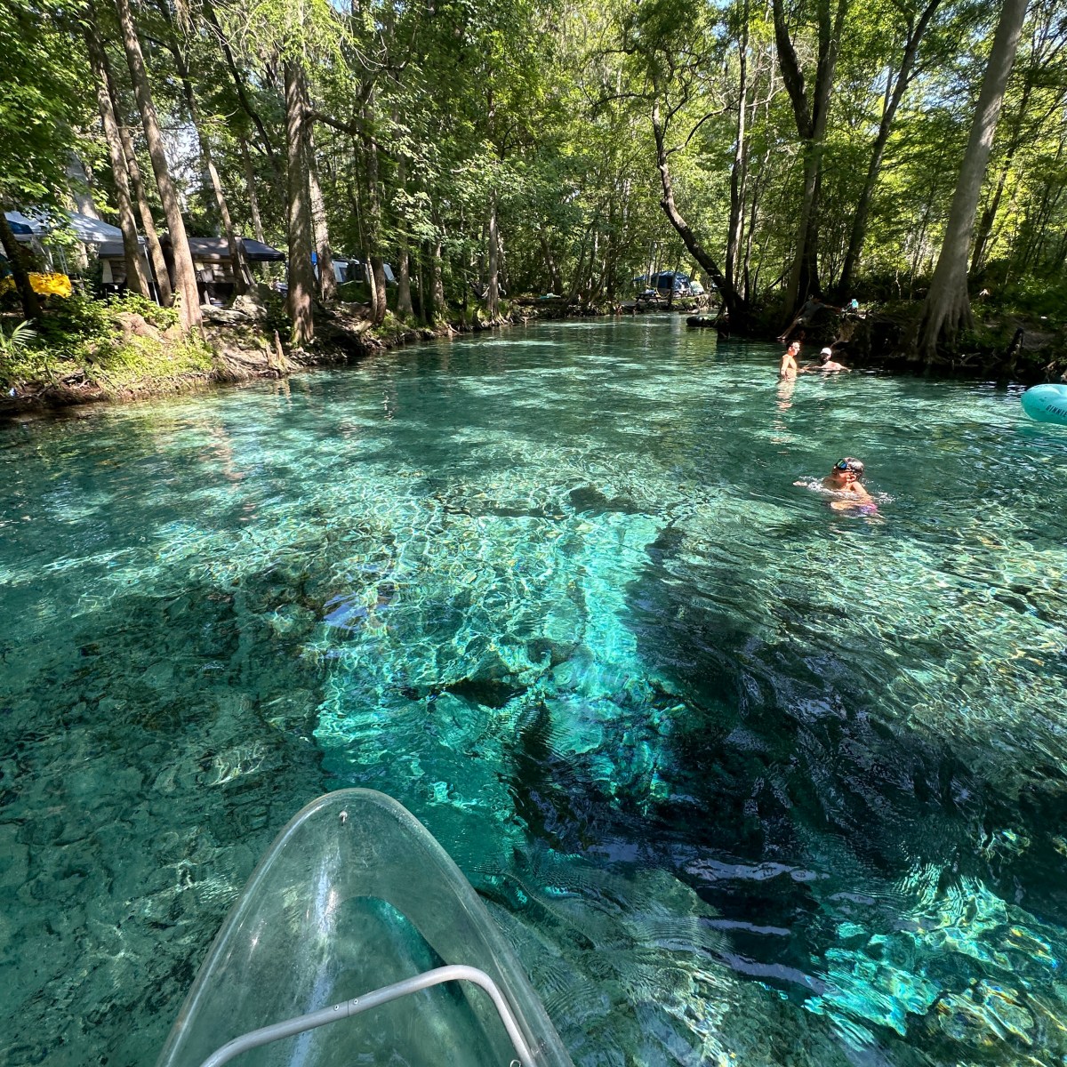 Clear kayak on a crystal-clear river with swimmers and lush green trees.